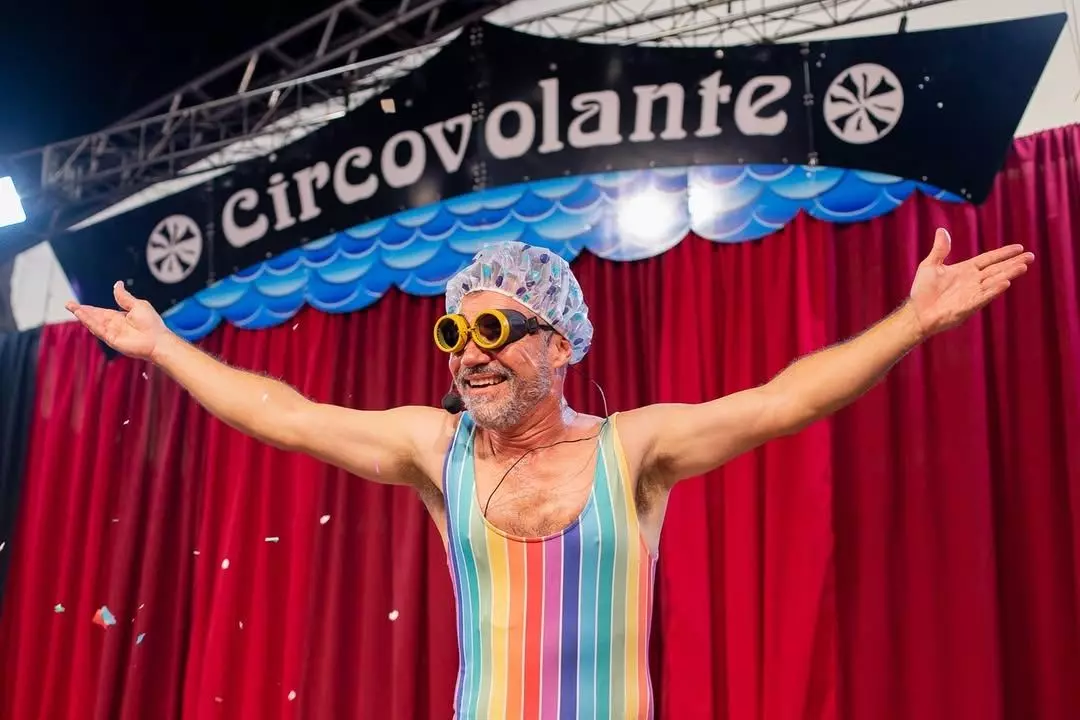 Palhaço do Circovolante com roupa listrada e óculos grandes se apresenta com os braços abertos no palco.