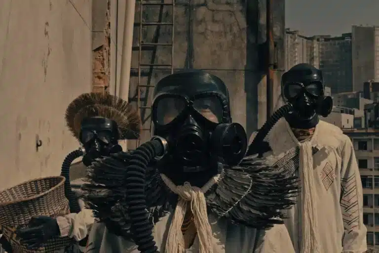 A imagem mostra três indígenas com trajes de proteção, como máscaras de gás. O da esquerda está com um cocar na cabeça, o do meio com o cocar pendurado no pescoço, a cena é parte do filme JAVYJU que será apresentado na mostra cine parentes