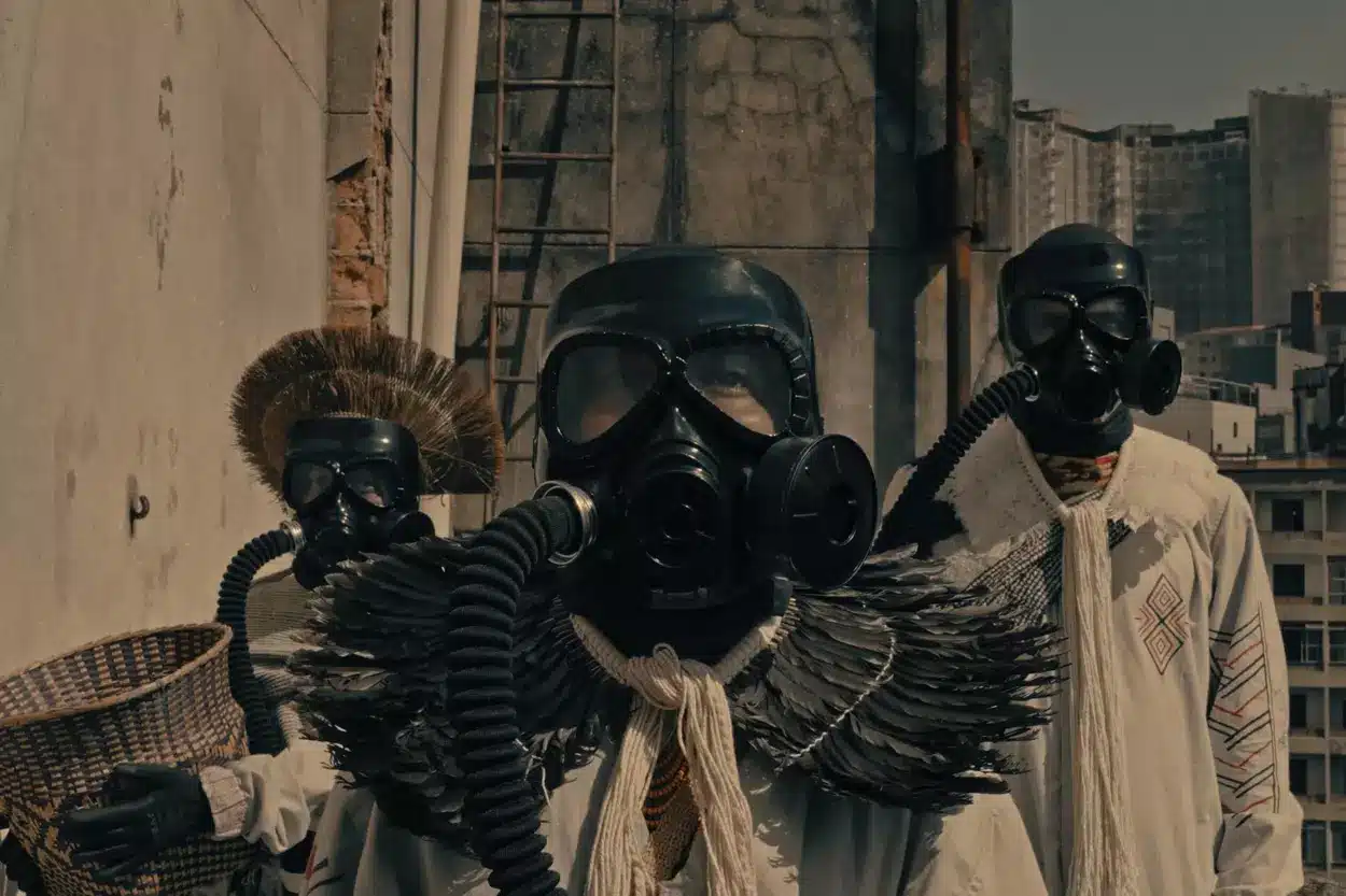 A imagem mostra três indígenas com trajes de proteção, como máscaras de gás. O da esquerda está com um cocar na cabeça, o do meio com o cocar pendurado no pescoço, a cena é parte do filme JAVYJU que será apresentado na mostra cine parentes