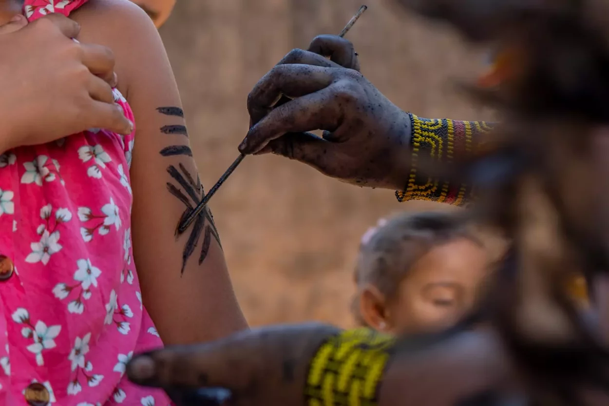 Mão de pessoa indígena com tinta escura fazendo uma pintura tradicional Borum-kren no braço de uma criança.