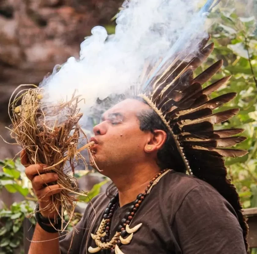 Danilo, cacique do povo indígena Borum-kren, soprando fumaça de um feixe de ervas secas acesas, em meio à natureza.