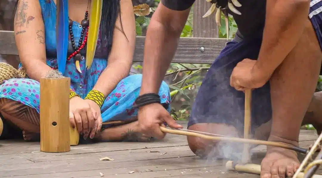Duas pessoas indígenas sentadas no chão de madeira, uma delas utilizando técnica tradicional de atrito com gravetos para fazer fogo, com fumaça subindo.