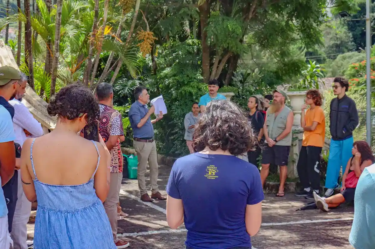 Grupo de estudantes reunidos ao ar livre, ouvindo um homem que segura papéis e fala diante de uma área com árvores na UFOP.