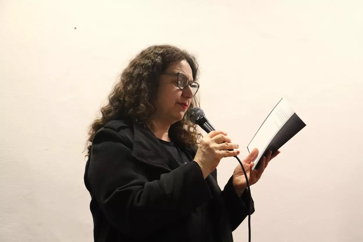 Guiomar, uma mulher de cabelos cacheados e óculos segura um microfone e lê atentamente um livro aberto. Ela veste casaco preto e está em pé diante de uma parede branca.