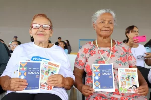Duas idosas seguram o Estatuto da Pessoa Idosa e gibi da Turma da Mônica sobre intergeracionalidade durante evento de lançamento.