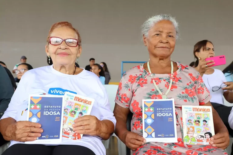 Duas idosas seguram o Estatuto da Pessoa Idosa e gibi da Turma da Mônica sobre intergeracionalidade durante evento de lançamento.
