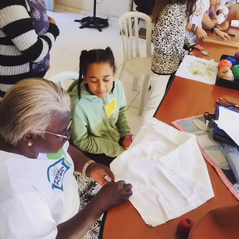 Idosa ensina menina a bordar tecido branco durante atividade intergeracional de artesanato.