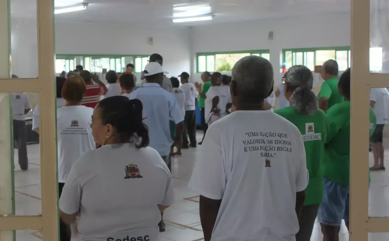 Idosos participam de atividade coletiva em salão, usando camisetas com frases de valorização da pessoa idosa.