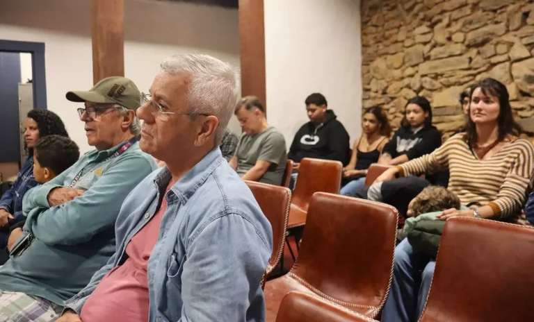 Foto de plateia sentada em cadeiras de couro marrom. Diversas pessoas, de diferentes idades, assistem ao evento. No fundo, uma parede de pedras compõe o ambiente. Algumas pessoas olham para frente com atenção, outras interagem entre si.