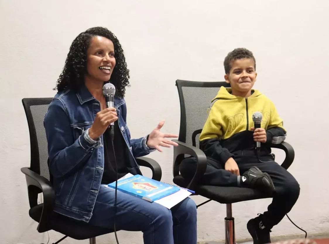 Em um ambiente interno com parede branca, Josiane, uma mulher de cabelos cacheados e jaqueta jeans segura um microfone enquanto fala sorrindo. Ao lado dela, um menino com moletom amarelo e preto, Théo, também com microfone na mão, sorri sentado em uma cadeira de escritório. Sobre o colo da mulher há um livro colorido.