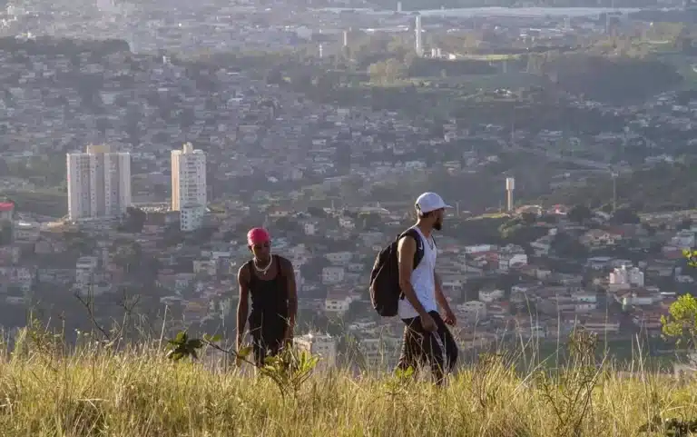 Dois jovens caminham em área de mata alta com vista panorâmica de cidade ao fundo, cena do curta que será exibido na Mostra Muito com Pouco.