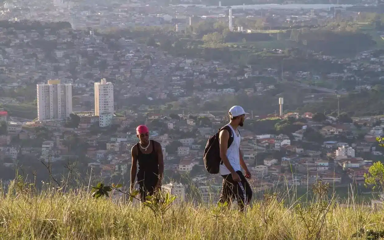 Dois jovens caminham em área de mata alta com vista panorâmica de cidade ao fundo, cena do curta que será exibido na Mostra Muito com Pouco.