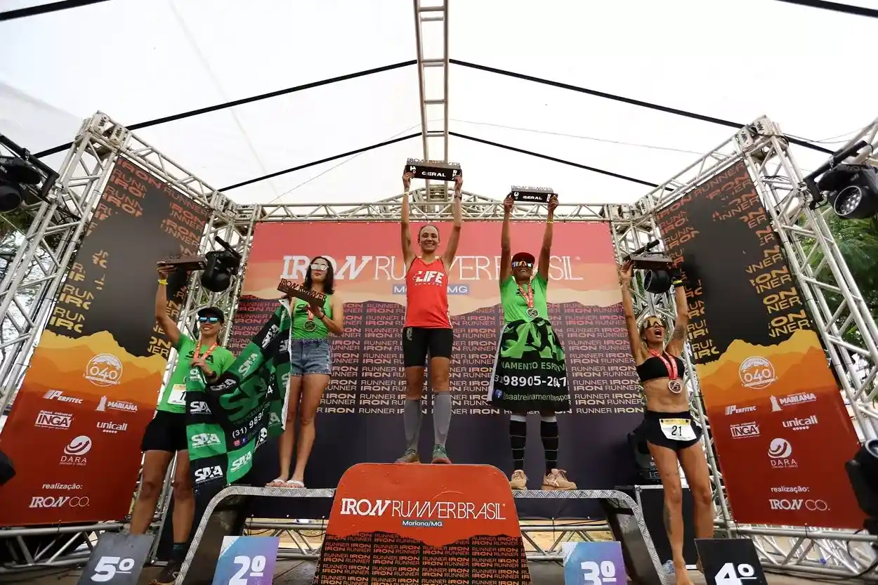 Mulheres celebram no pódio do Iron Runner em Mariana, segurando troféus e medalhas.