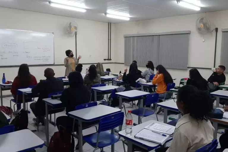 Inscrições abertas PREMAR Minas Gerais. Professora diante de quadro branco, explicando a alunos, em suas carteiras, detalhes sobre redação.