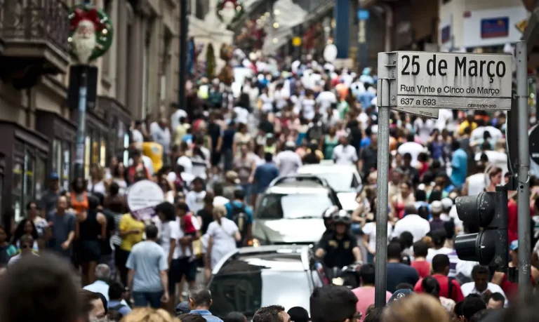 Multidão caminha pela Rua 25 de Março, em São Paulo, com lojas e carros ao redor. Indicadores como renda e acesso a serviços nas áreas urbanas influenciam o desempenho do Brasil no IDH.