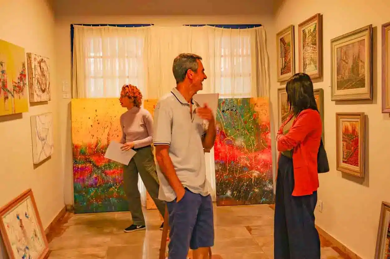 Na imagem é possível ver pessoas apreciando obras de arte numa sala de exposição do Ateliê Aberto.
