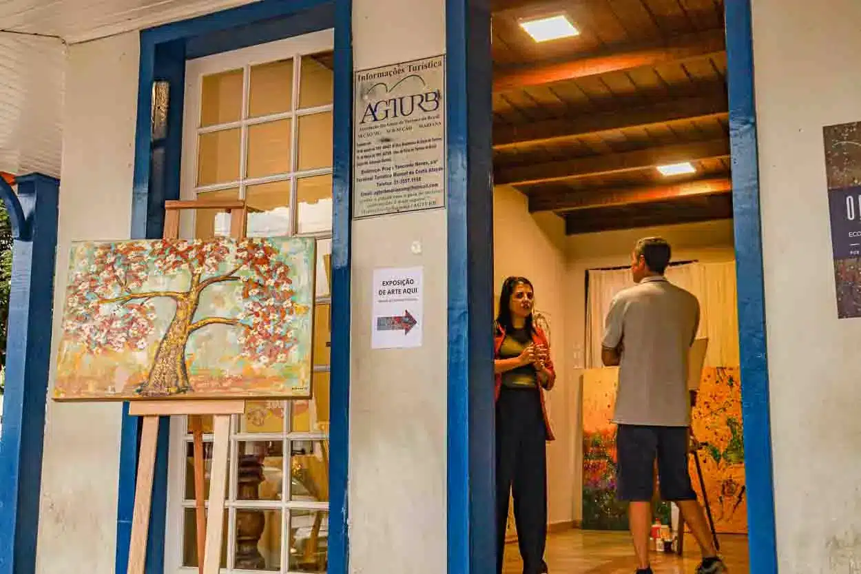 Na imagem se vê uma sala, com o artista e uma visitante. Também há um quadro ao lado de fora da porta e uma placa convidando as pessoas a visitarem a exposição de arte. Na sala acontecerá o projeto Ateliê Aberto.