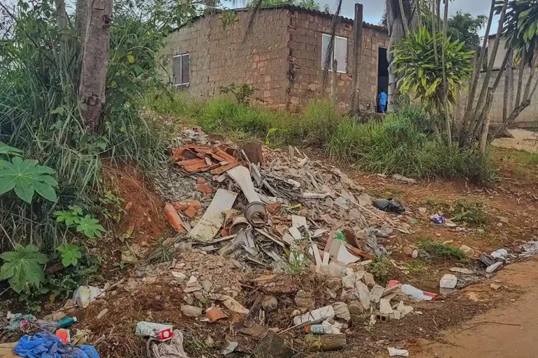 Montanha de entulho com tijolos, madeira e lixo espalhado em frente a uma casa deteriorada.