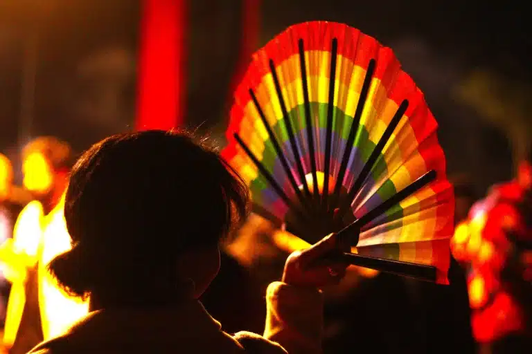 A foto mostra uma mulher segurando um leque com a bandeira LGBT+ durante a parada.