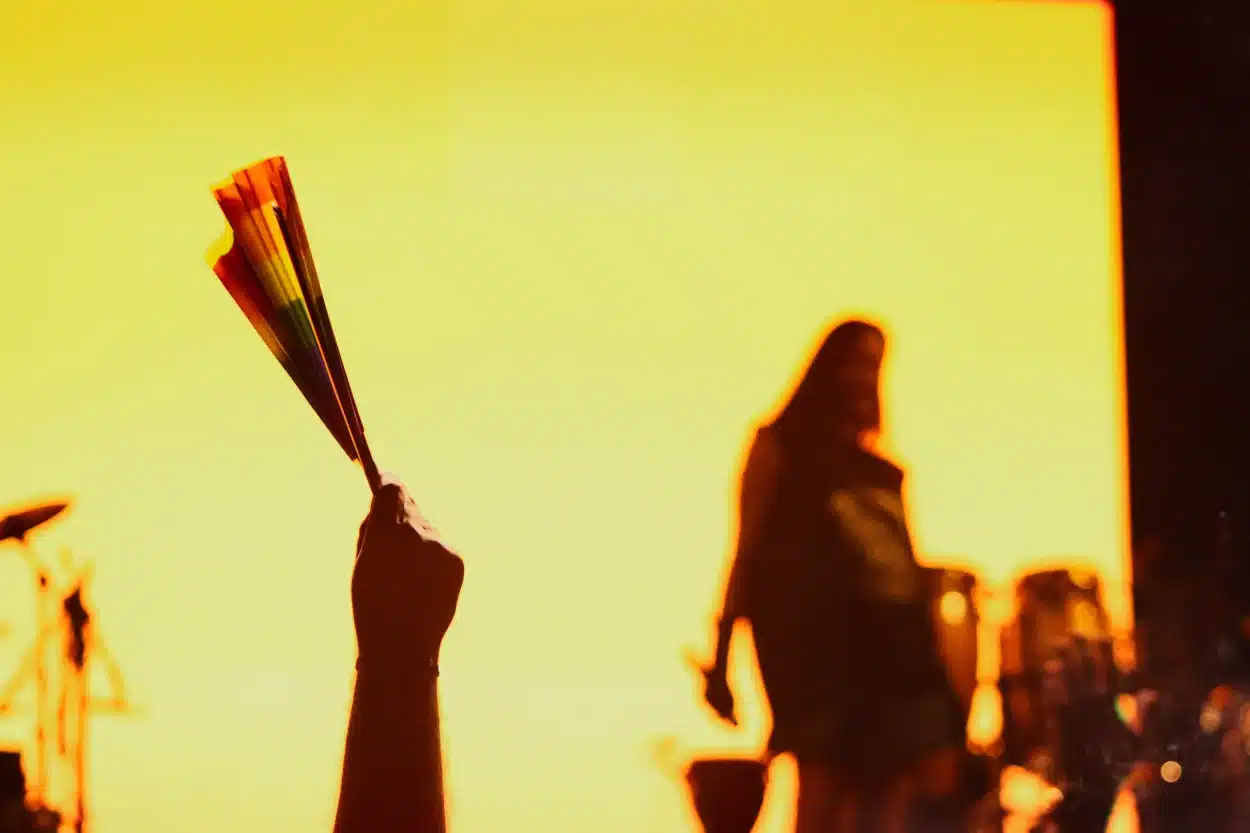 A foto mostra um leque com as cores da bandeira lgbt+ e a cantora Jaloo ao fundo
