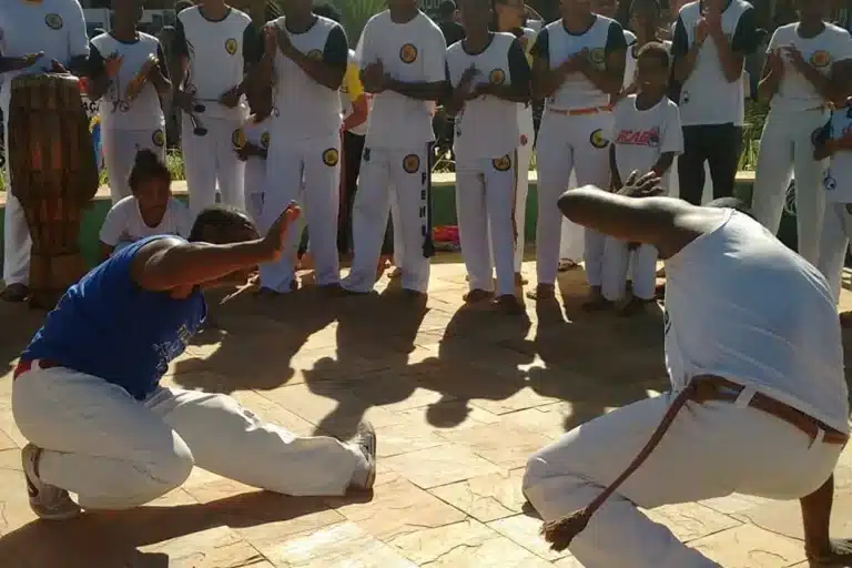 Dois capoeiristas em movimento dentro de uma roda de capoeira ao ar livre.