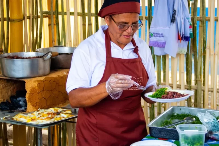 Mulher quitandeira, usando avental bordô e touca preta com detalhe vermelho, serve prato com comida típica mineira durante o Festival do Salto.