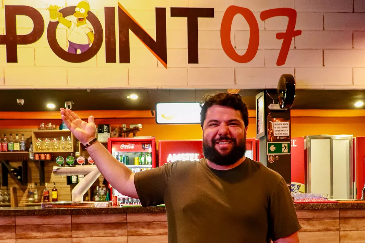 O sócio do Point 07, Aquiles, sorri para a câmera e aponta para o balcão onde está escrito Point 07