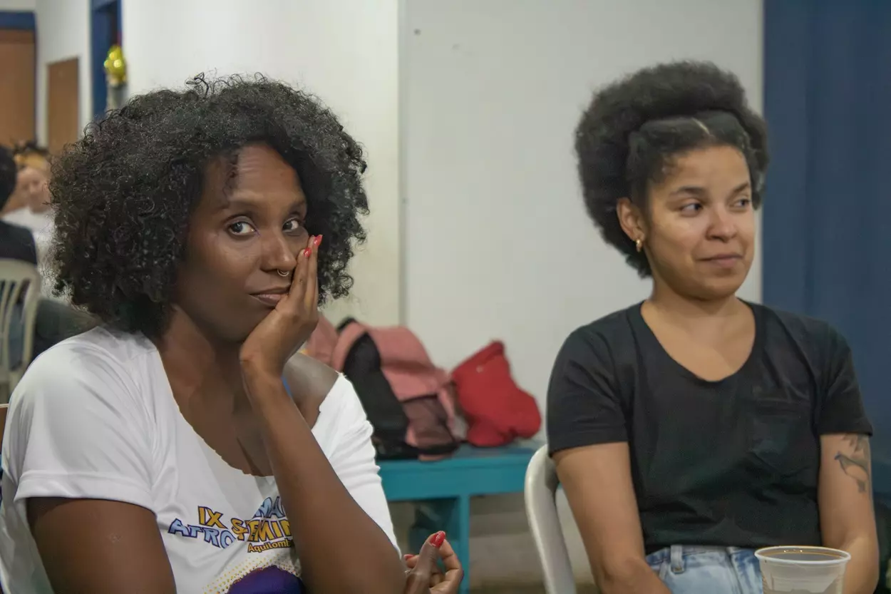 Duas mulheres Negras, Uma delas, à esquerda, Dani olha para a câmera e mantêm uma de suas mãos no queixo, a expressão é serena