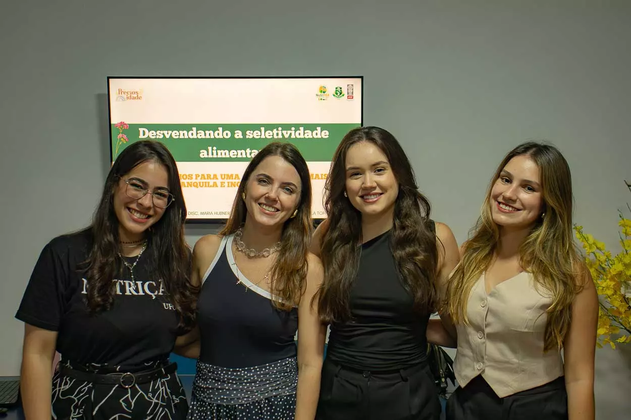 Quatro mulheres sorriem e posam para a câmera em frente uma TV onde a gente vê a seguinte frase: "Desvendando a seletividade alimentar"
