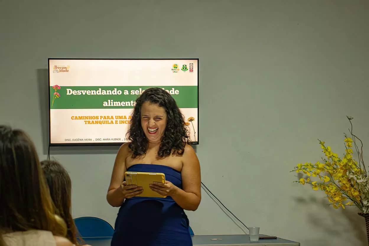 Uma mulher em frente a uma TV com um tablet na mão sorri com muito entusiasmo, atrás dela tem ainda uma mesa e ao lado um vaso com flores amarelas. Ela parece discursar para outras mulheres em uma sala