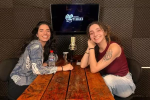 duas mulheres sorriem para a câmera, elas estão sentadas uma de frente para a outra, uma mesa as separa, ao fundo uma tv exibe a logo da Entrevista Primaz e sobre a mesa tem um tablet, um microfone e algumas canecas