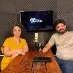 Fotografia em plano médio de duas pessoas sentadas no estúdio de gravação da Agência Primaz. À esquerda, Luciane Trevisan, uma mulher branca de cabelos escuros e cacheados, veste uma blusa amarela e sorri suavemente para a câmara. À direita, o entrevistador, Lui Pereira, um homem branco com barba e cabelos pretos curtos, veste uma camiseta cinzenta e calça jeans azul. Eles estão sentados frente a frente numa mesa comprida de madeira rústica. No centro da mesa, encontra-se um microfone de condensador prateado de estilo vintage e duas canecas. Ao fundo, a parede é revestida com painéis de espuma acústica cinza-escura. Entre os dois, há uma TV que exibe o logótipo do programa "Entrevista Primaz" em letras brancas sobre um fundo escuro. A iluminação é quente e o ambiente é de uma conversa de podcast.