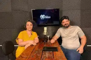 Fotografia em plano médio de duas pessoas sentadas no estúdio de gravação da Agência Primaz. À esquerda, Luciane Trevisan, uma mulher branca de cabelos escuros e cacheados, veste uma blusa amarela e sorri suavemente para a câmara. À direita, o entrevistador, Lui Pereira, um homem branco com barba e cabelos pretos curtos, veste uma camiseta cinzenta e calça jeans azul. Eles estão sentados frente a frente numa mesa comprida de madeira rústica. No centro da mesa, encontra-se um microfone de condensador prateado de estilo vintage e duas canecas. Ao fundo, a parede é revestida com painéis de espuma acústica cinza-escura. Entre os dois, há uma TV que exibe o logótipo do programa "Entrevista Primaz" em letras brancas sobre um fundo escuro. A iluminação é quente e o ambiente é de uma conversa de podcast.