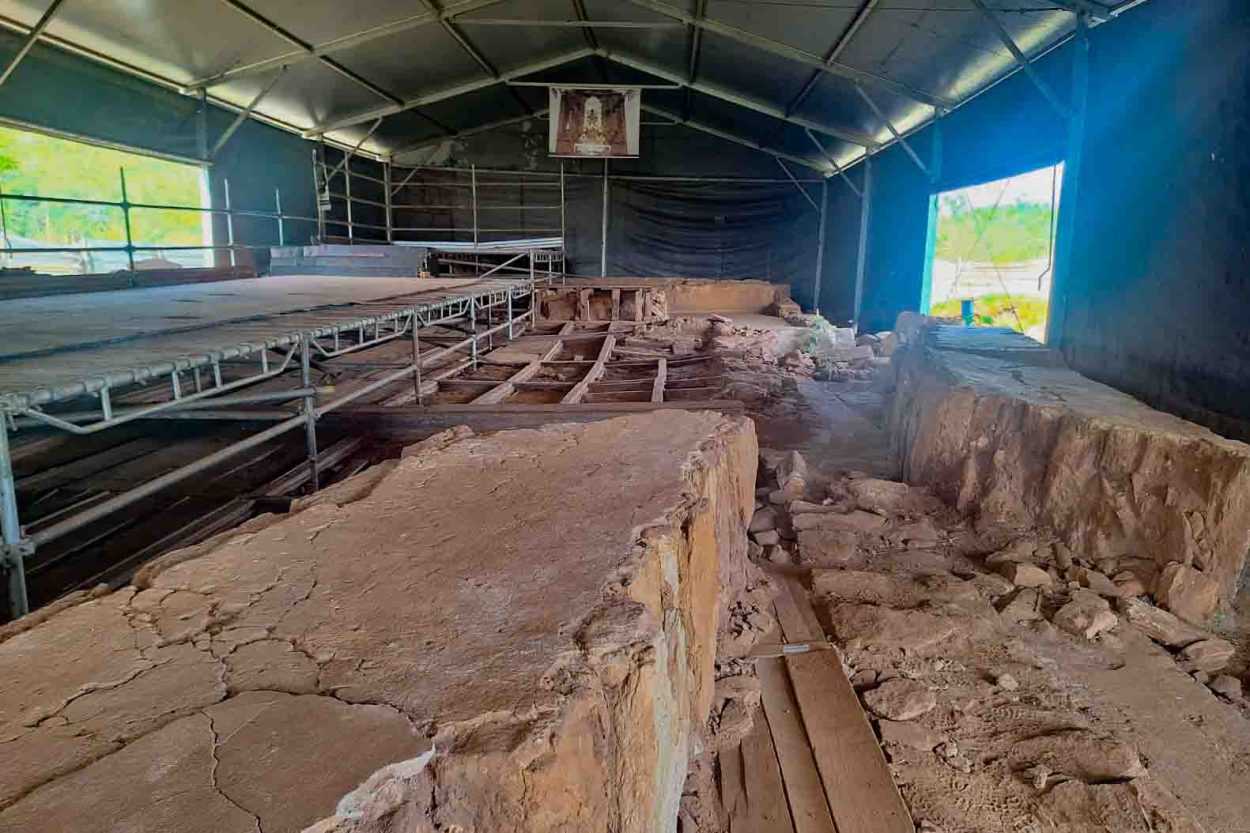 Uma fotografia do interior das ruínas da Igreja de São Bento, em Bento Rodrigues (Mariana, MG). As paredes remanescentes, feitas de barro e pedras em tons terrosos e ocres, estão severamente desgastadas e fragmentadas. Elas são protegidas por uma grande estrutura metálica moderna que funciona como um galpão temporário, com telhado de zinco e andaimes de aço ao redor das ruínas para estabilização. Ao fundo, uma pequena imagem religiosa está pendurada no topo da estrutura. A luz natural entra pelas laterais abertas do galpão, iluminando o solo irregular e os escombros preservados.