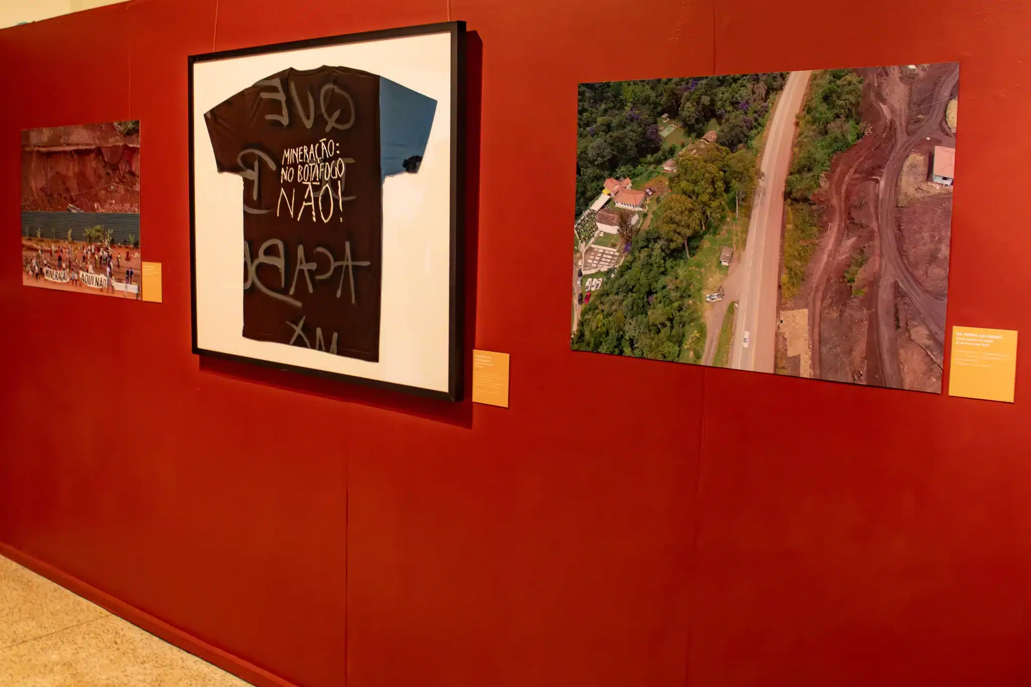 A imagem mostra uma parede vermelha de uma galeria com três obras de arte emolduradas e etiquetas informativas amarelas ao lado de cada uma. À esquerda, uma fotografia pequena mostra um grupo de pessoas com uma faixa MINERAÇÃO AQUI NÃO em frente a uma área de terra escavada. No centro, uma camiseta preta grande emoldurada tem texto branco pintado à mão: MINERAÇÃO NO BOTAFOGO NÃO! ACABA, com algumas palavras invertidas. À direita, uma fotografia aérea maior mostra uma estrada dividindo uma paisagem: um lado com vegetação verde e casas, e o outro com terra vermelha e marcas de mineração. O chão é de pedra clara.