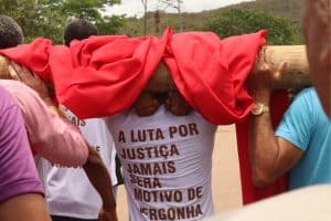 A foto, tirada de costas em um ângulo médio, mostra um grupo de pessoas carregando coletivamente um grande tronco de madeira sobre os ombros. O tronco está parcialmente coberto por um tecido vermelho vibrante que cai pelas laterais. No centro da imagem, um homem veste uma camiseta branca com uma frase de protesto escrita em letras maiúsculas de cor marrom: "A LUTA POR JUSTIÇA JAMAIS SERÁ MOTIVO DE VERGONHA" Detalhes da cena: Composição: Outras pessoas aparecem nas bordas da foto, ajudando a sustentar o peso do tronco. À direita, vê-se o braço de um homem com um relógio dourado e uma camiseta azul; à esquerda, parte de uma camiseta rosa e outra branca. Ambiente: O cenário ao fundo é ao ar livre, mostrando uma estrada de terra ou asfalto claro, cercada por árvores verdes e morros sob um céu nublado. Atmosfera: A imagem transmite um sentimento de esforço coletivo, resistência e mobilização social.