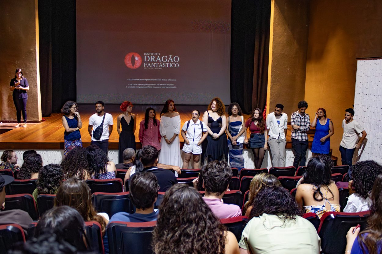 Uma foto horizontal, de plano médio, mostra um grupo diverso de treze pessoas, homens e mulheres, em pé em um palco de teatro, de frente para uma plateia lotada. Eles estão alinhados em frente a uma grande tela de projeção que exibe o logotipo vermelho e branco do "Instituto Dragão Fantástico" e texto de direitos autorais. Uma mulher na extrema esquerda do palco segura um microfone e gesticula enquanto fala. As pessoas no palco usam uma variedade de roupas casuais e formais. A plateia, vista das fileiras de trás, preenche o teatro e está voltada para o palco, com as costas das cabeças visíveis. O teatro tem paredes escuras e cortinas pretas ao lado da tela. A iluminação é focada no palco.